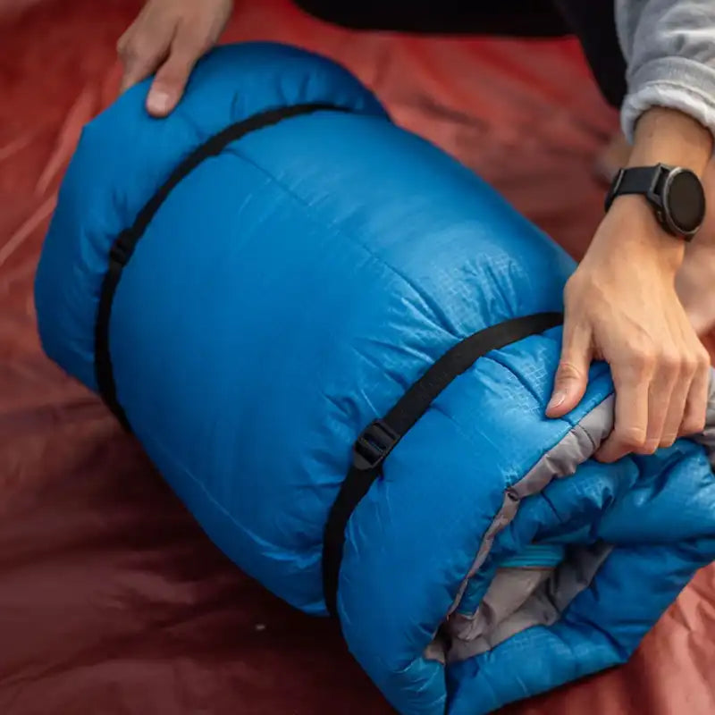 A person's hands are shown rolling up a bright blue sleeping bag owith black straps