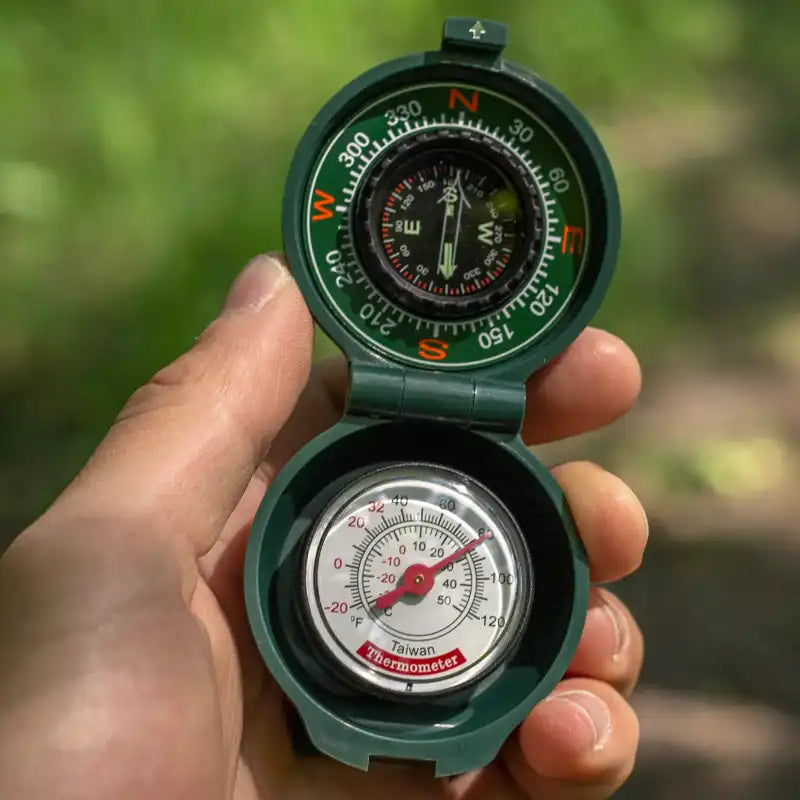 A green folding pocket compass with an integrated thermometer, held in a person's hand against a blurred green outdoor background.